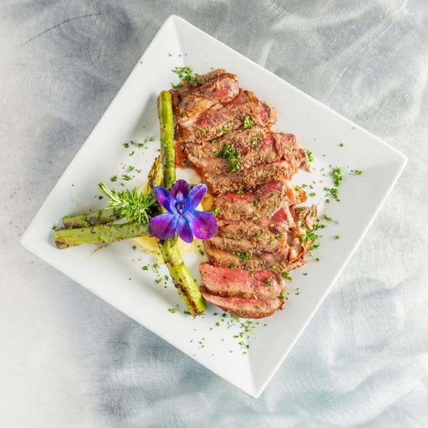 Sliced steak with asparagus, mashed potatoes, rosemary, and a purple orchid on a white plate.