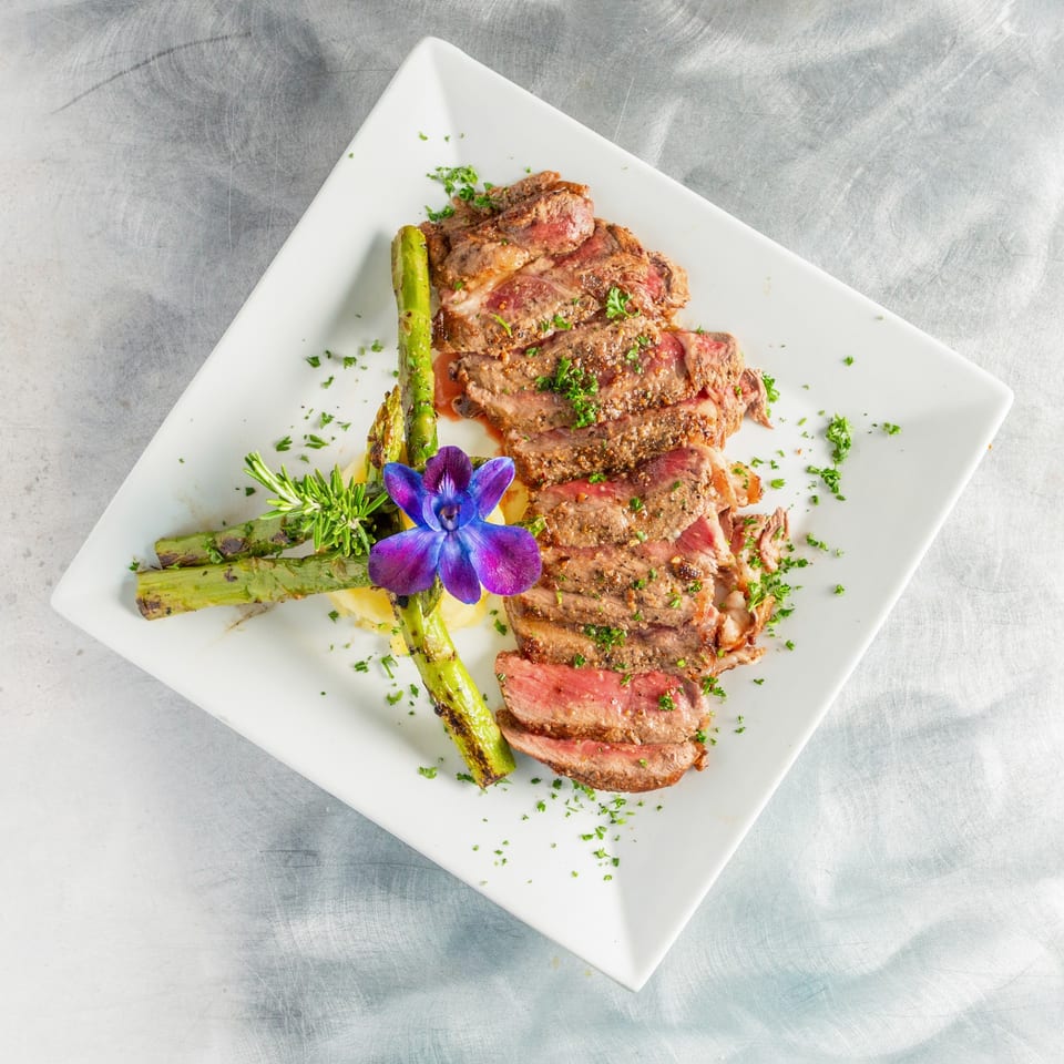 Sliced steak with asparagus, mashed potatoes, rosemary, and a purple orchid on a white plate.