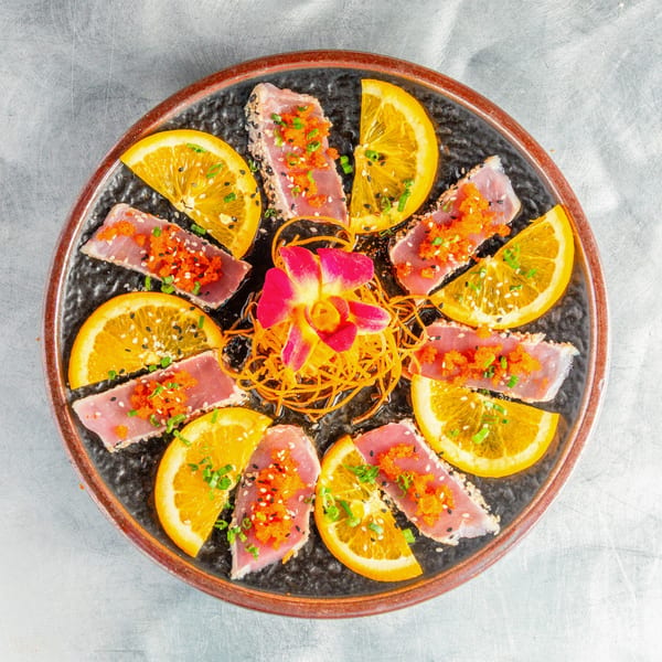 Seared tuna sashimi with orange slices, tobiko, sesame seeds, and a pink orchid on a dark plate.