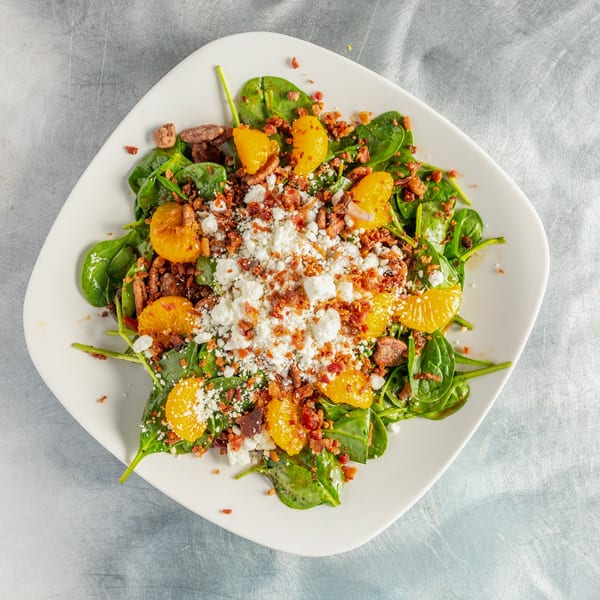 Spinach salad with mandarin oranges, crumbled bacon, and feta cheese on a white plate.